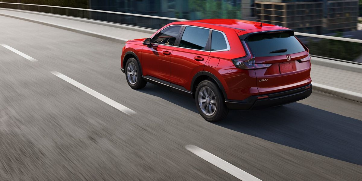 2027 Honda CR-V EX in Radiant Red Metallic back driving on a city bridge through the downtown skyline (Compact Crossover SUV).